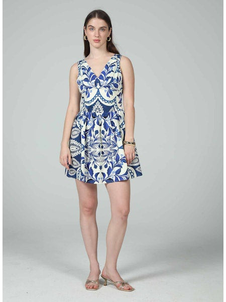 A woman stands against a plain gray background, wearing the Zertz ELIANA TIE MINI DRESS—a sleeveless V-neck mini dress with a blue and white floral pattern. She pairs it with hoop earrings, sandals, and a bracelet.