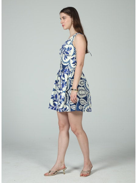 A woman in profile wears the Zertz ELIANA TIE MINI DRESS—sleeveless, blue with white florals—and beige heeled sandals. She has long brown hair, a neutral expression, and stands against a plain light gray background.
