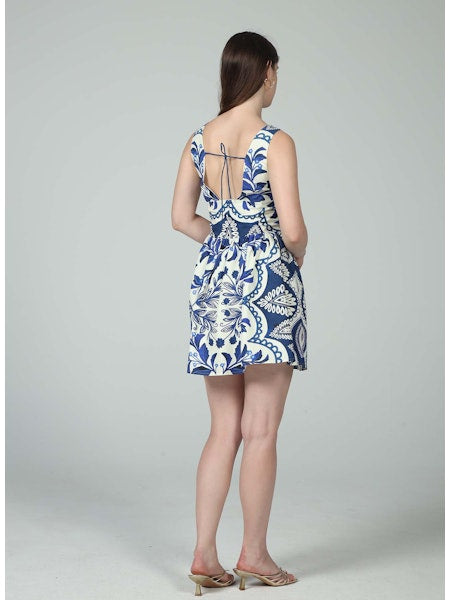 A woman faces away in the Zertz ELIANA TIE MINI DRESS, featuring a blue hue, low back, thin tie straps, and nude heeled sandals. Her long brown hair cascades down her back against a plain light gray background.