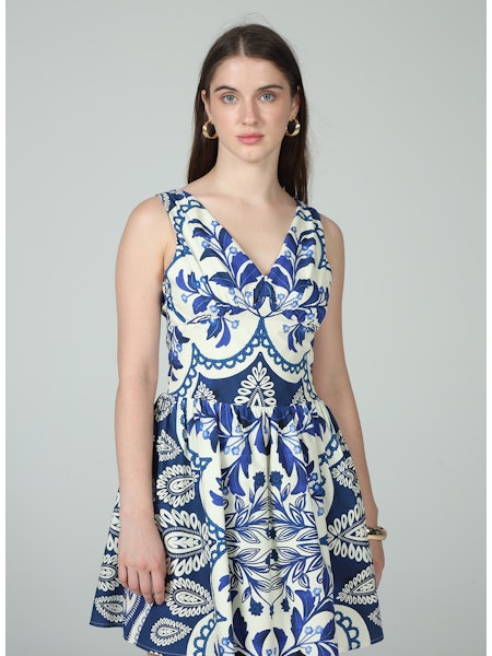 A woman with long brown hair wears gold hoop earrings and the Zertz ELIANA TIE MINI DRESS, a sleeveless blue-and-white patterned mini dress, standing against a plain light gray background and gazing slightly to the side.
