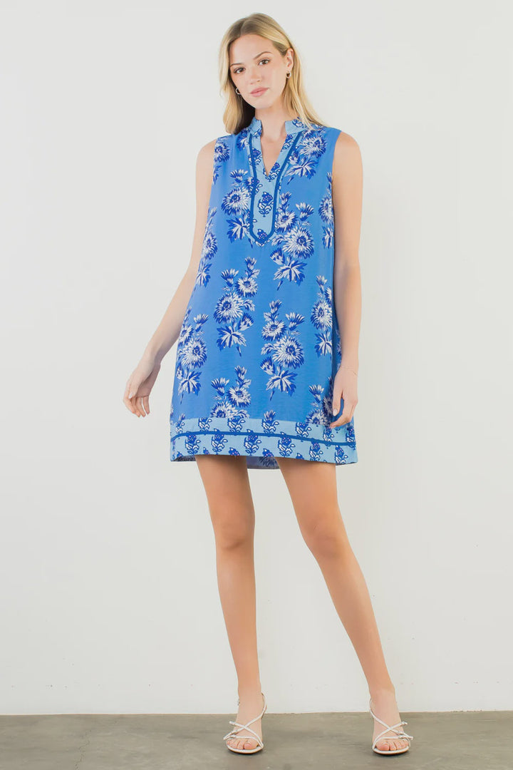 A woman with light hair and a neutral expression wears the Zertz SLEEVELESS FLORAL PRINT DRESS—a V-neck, summer-ready style—against a white wall, paired with white sandals and one leg slightly bent.