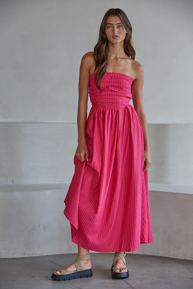A woman with long brown hair wears the Zertz PROMENADE DRESS L6998, a strapless bright pink pleated midi dress, styled with beige platform sandals. She stands indoors against a plain light gray background, looking at the camera.