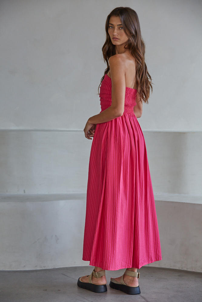 A woman with long brown hair models the Zertz PROMENADE DRESS L6998, a strapless, ankle-length bright pink gown with a smocked bodice and pleated skirt. She stands sideways indoors, glancing over her shoulder.