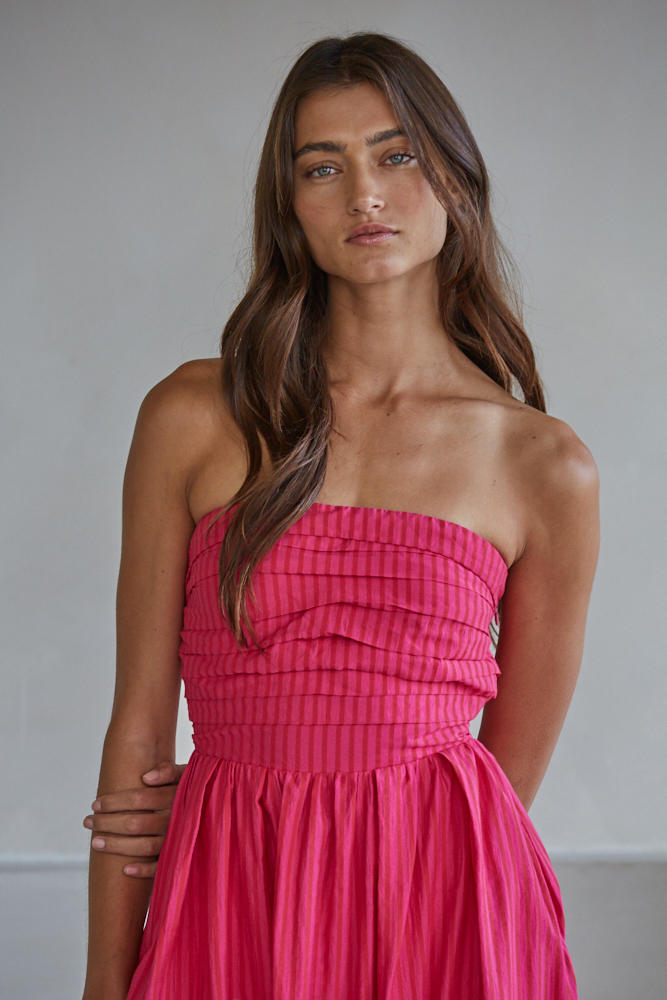 A woman with long brown hair wears the Zertz PROMENADE DRESS L6998, a strapless, pleated, bright pink dress, standing against a plain light background and looking at the camera with a neutral expression.