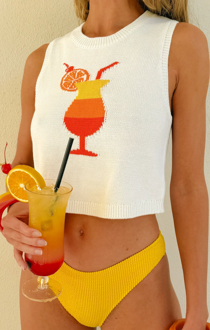 Wearing the Zertz MILO TANK, a white cropped knit top with a fruity cocktail graphic, and yellow textured bikini bottoms, she holds a tropical drink—ideal for relaxed beachside lounging.