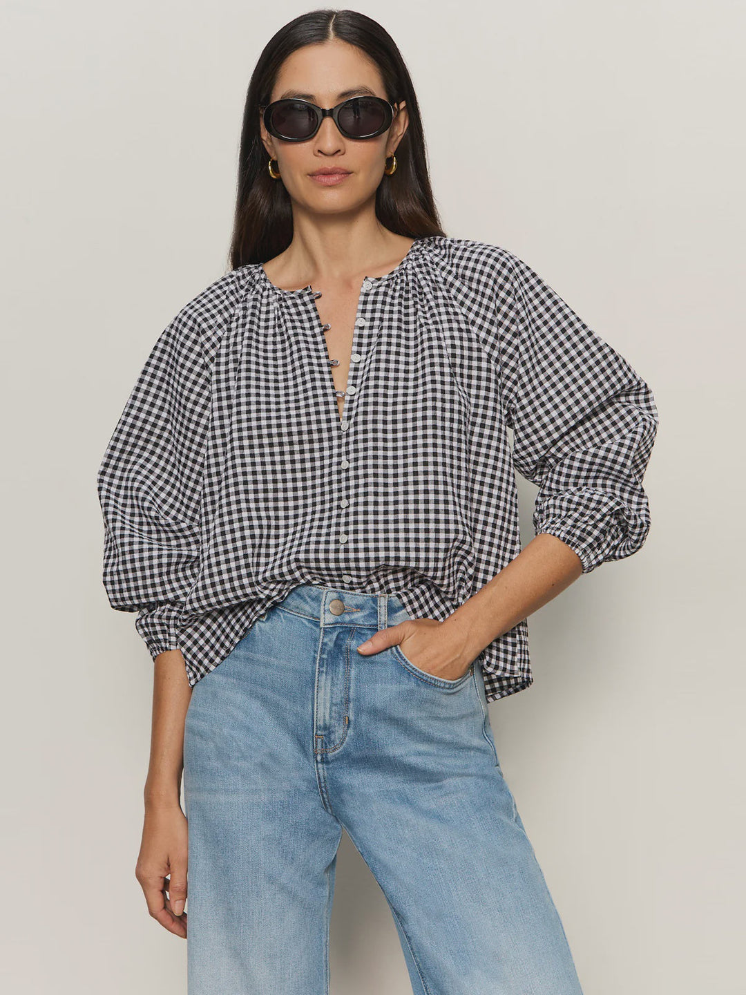 A woman wearing black sunglasses, light blue high-waisted jeans, and the Zertz PERFECT GINGHAM TOP—a loose black-and-white checked blouse with raglan sleeves—stands against a neutral background with one hand in her pocket.