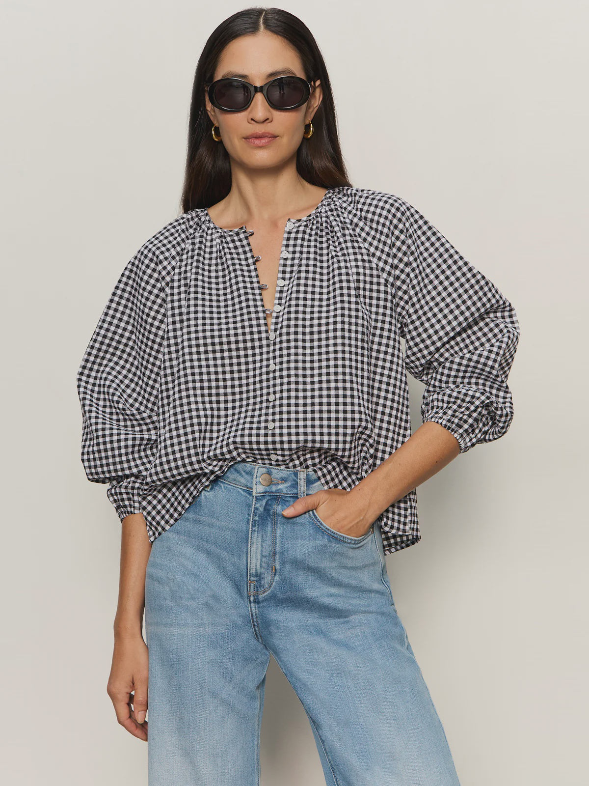 A woman wearing black sunglasses, light blue high-waisted jeans, and the Zertz PERFECT GINGHAM TOP—a loose black-and-white checked blouse with raglan sleeves—stands against a neutral background with one hand in her pocket.
