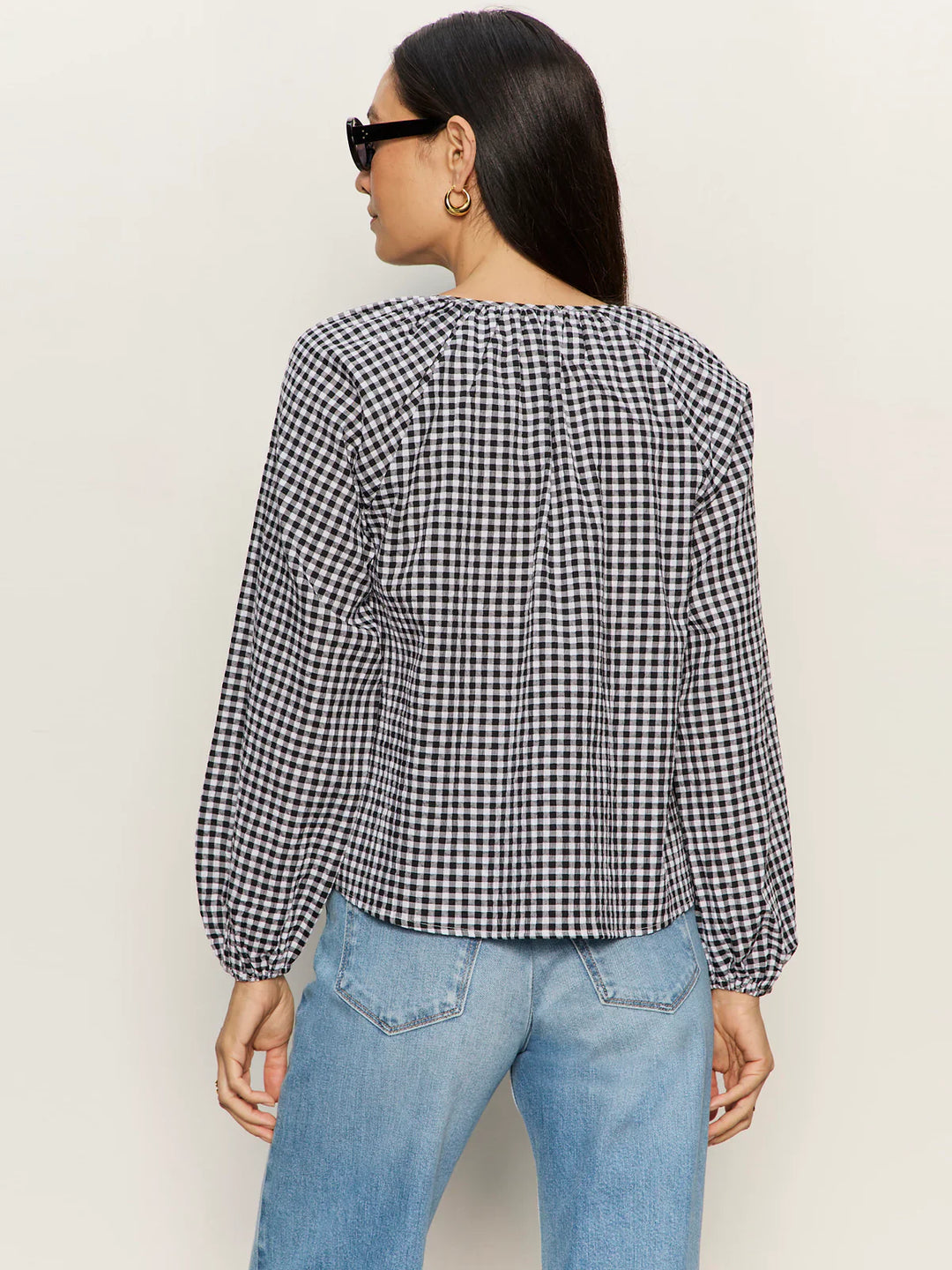 A woman with long dark hair, gold hoop earrings, and sunglasses models the Zertz PERFECT GINGHAM TOP—a relaxed-fit black and white checkered blouse—with light blue jeans, standing with her back to the camera against a plain backdrop.