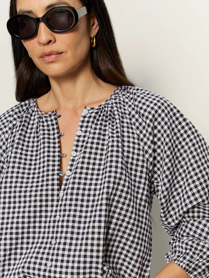 A woman wearing black sunglasses, gold hoop earrings, and the Zertz PERFECT GINGHAM TOP—a lightweight black-and-white checkered blouse with a split neck—stands against a plain light background.