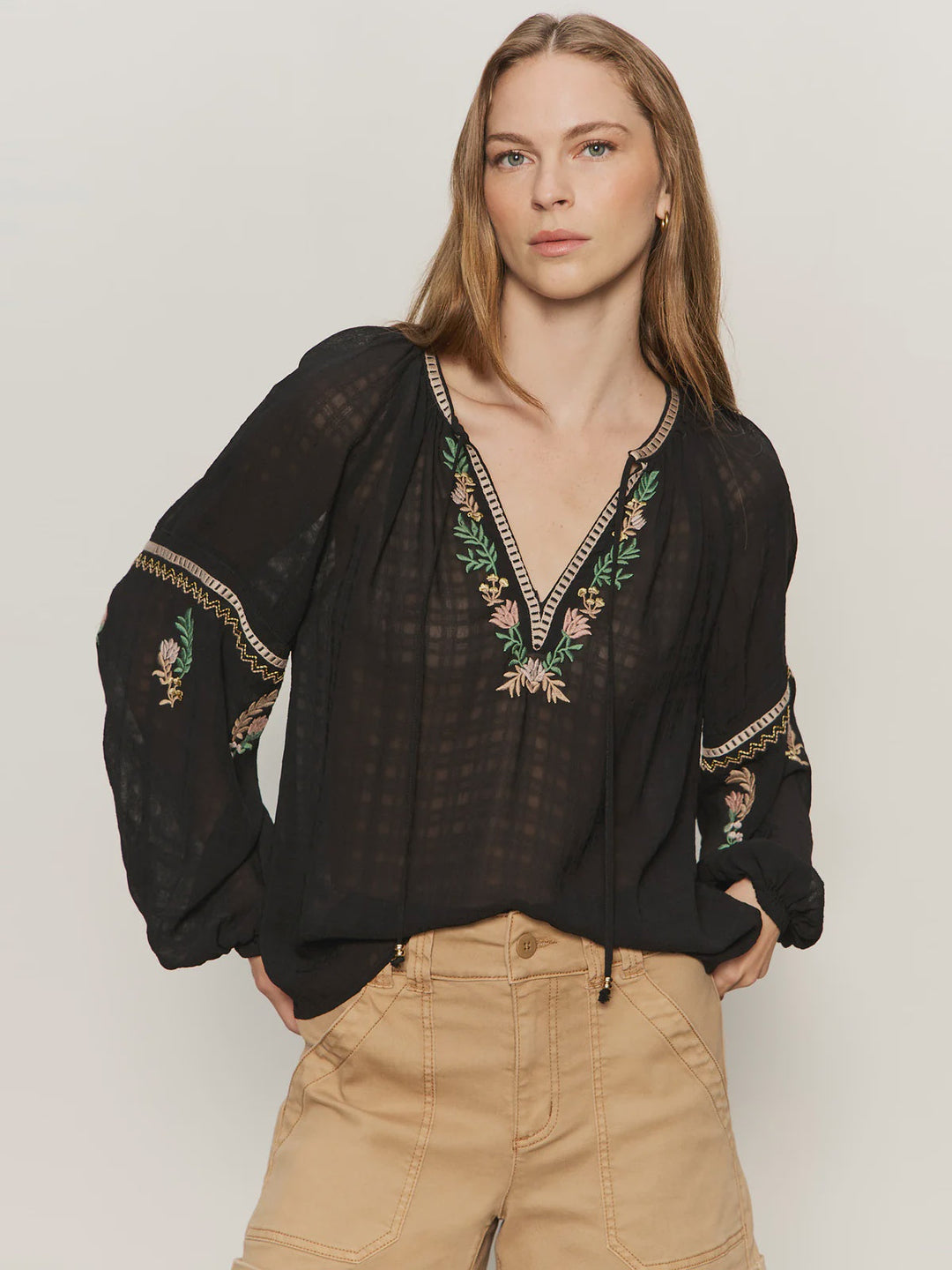 A woman with long, light brown hair wears the Zertz RAYE EMBROIDERED BLOUSE, a black semi-sheer top with a tie neck and floral embroidery, paired with tan high-waisted shorts against a plain light background.