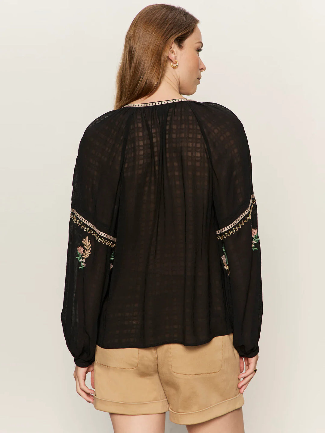 A woman with long brown hair is seen from behind, wearing the Zertz RAYE EMBROIDERED BLOUSE—a sheer black, lightweight top with floral embroidery on billowy sleeves—paired effortlessly with beige shorts.