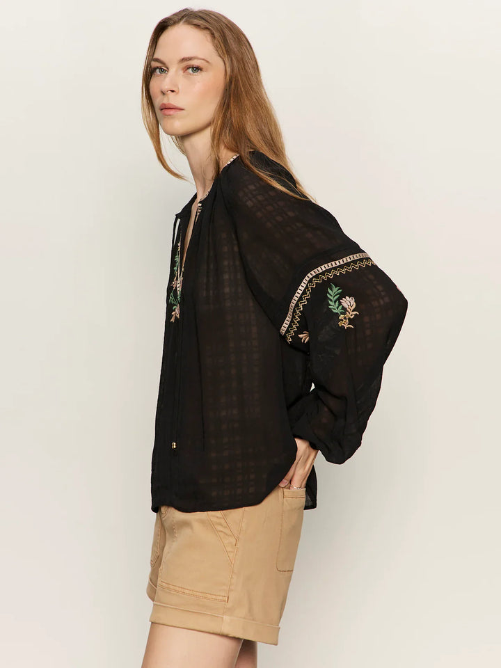 A woman with long, light brown hair wears the Zertz RAYE EMBROIDERED BLOUSE—a lightweight, sheer black top featuring floral embroidery on the sleeves—paired with tan shorts against a plain, light-colored background.