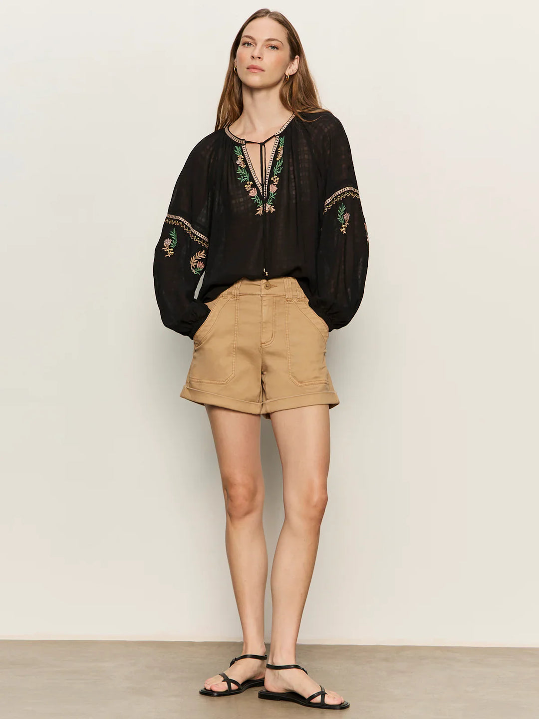 A woman stands against a plain background in the Zertz RAYE EMBROIDERED BLOUSE, tan high-waisted shorts, and black sandals. Her hair is down and she gazes slightly to the side with a neutral expression.