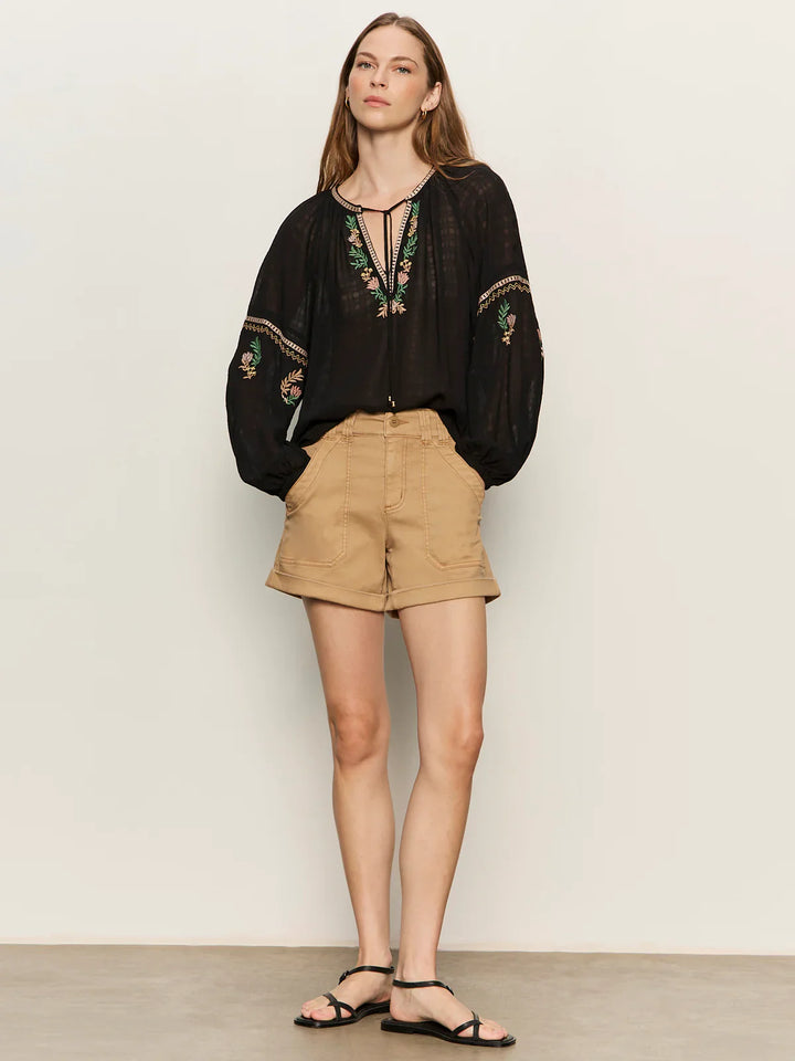 A woman stands against a plain background in the Zertz RAYE EMBROIDERED BLOUSE, tan high-waisted shorts, and black sandals. Her hair is down and she gazes slightly to the side with a neutral expression.