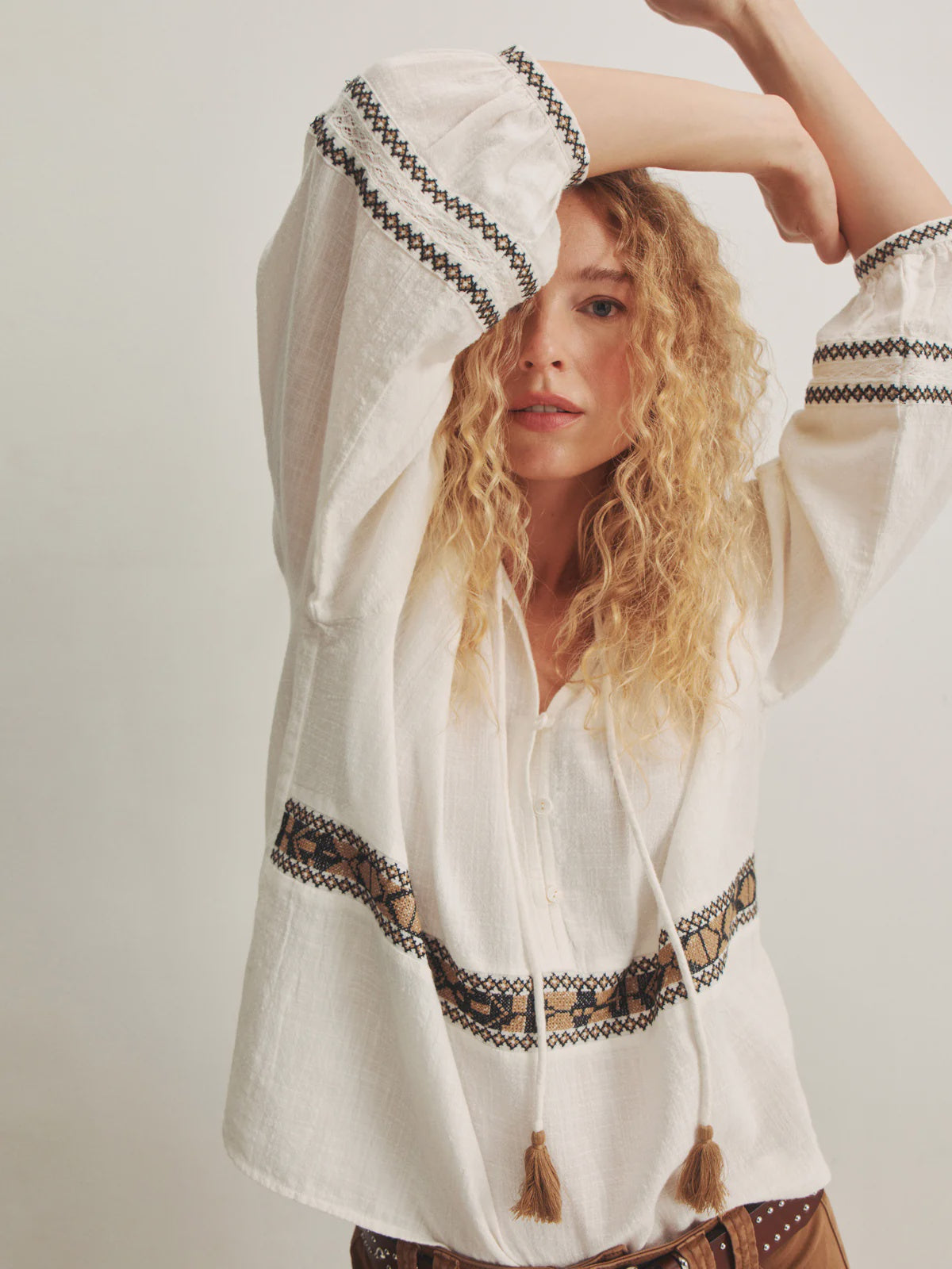 A woman with curly blonde hair poses with one arm raised, wearing the Zertz RETREAT EMB BLOUSE featuring embroidered details and tassels, paired with brown pants and a studded belt, against a plain light background.