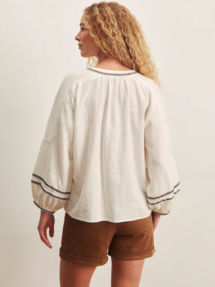 A woman with curly blonde hair is seen from the back wearing the Zertz RETREAT EMB BLOUSE, a loose-fitting embroidered top with decorative trim at the cuffs and neckline, paired with brown shorts against a plain light background.
