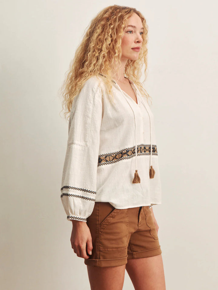 A woman with long curly blonde hair stands sideways against a plain light background, wearing the Zertz RETREAT EMB BLOUSE—an embroidered V-neck style with tassels and decorative trim—paired with brown shorts.