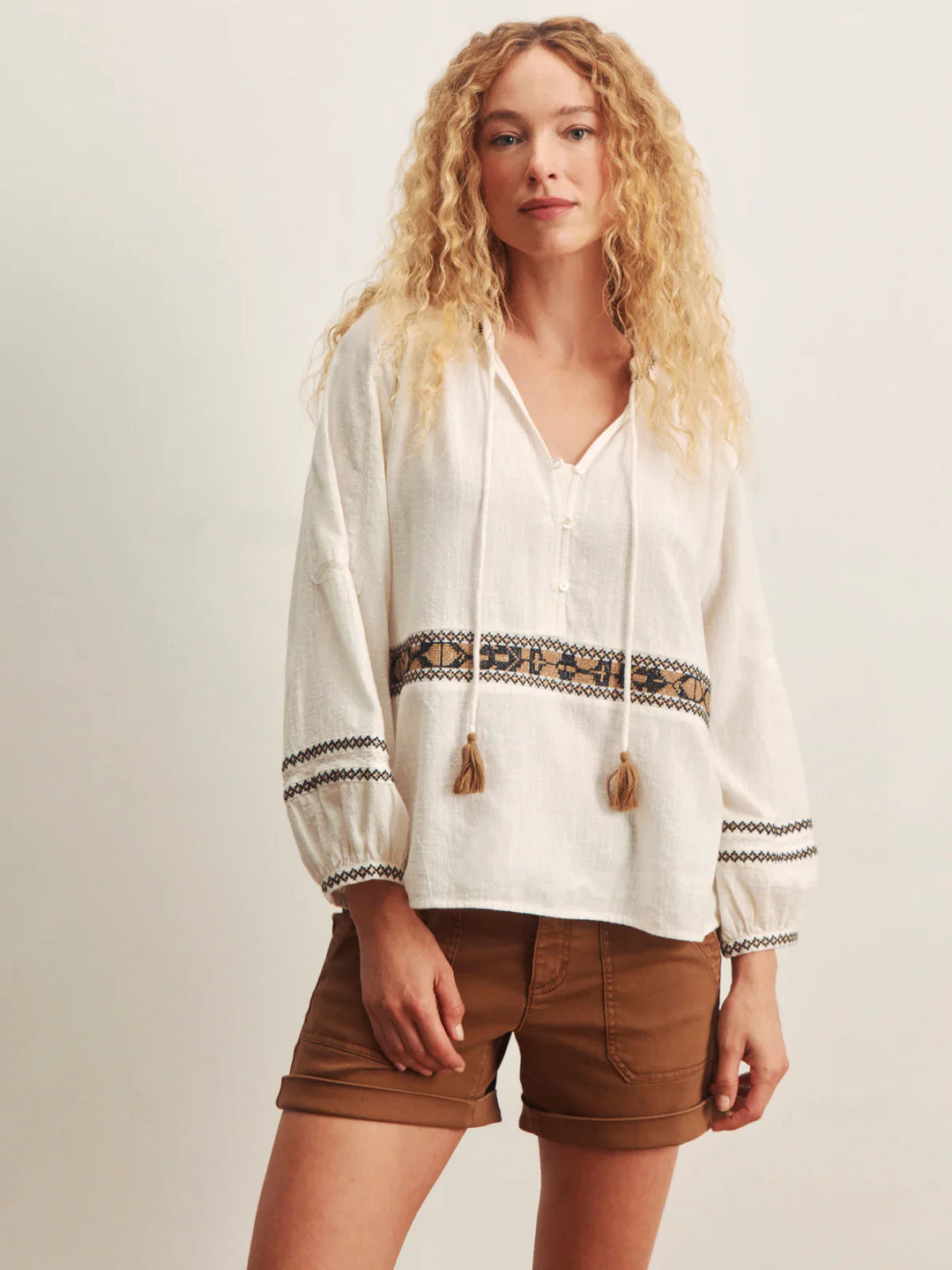 A woman with long curly blond hair models the Zertz RETREAT EMB BLOUSE, featuring embroidery and tassels, paired with brown shorts against a neutral background.