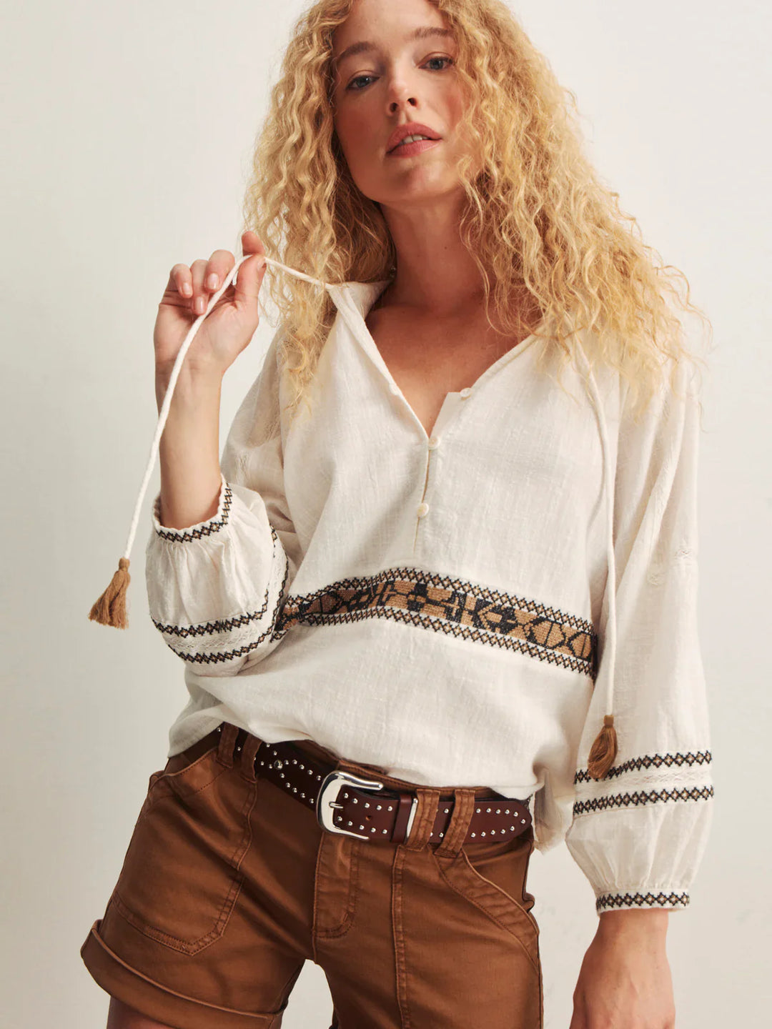 A woman with long curly blonde hair wears the Zertz RETREAT EMB BLOUSE, a loose embroidered V-neck top, paired with brown shorts and a studded belt. She holds one blouse tie, standing against a plain background.