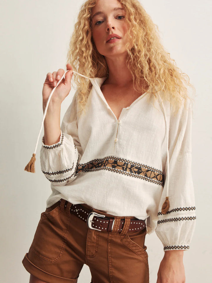 A woman with long curly blonde hair wears the Zertz RETREAT EMB BLOUSE, a loose embroidered V-neck top, paired with brown shorts and a studded belt. She holds one blouse tie, standing against a plain background.