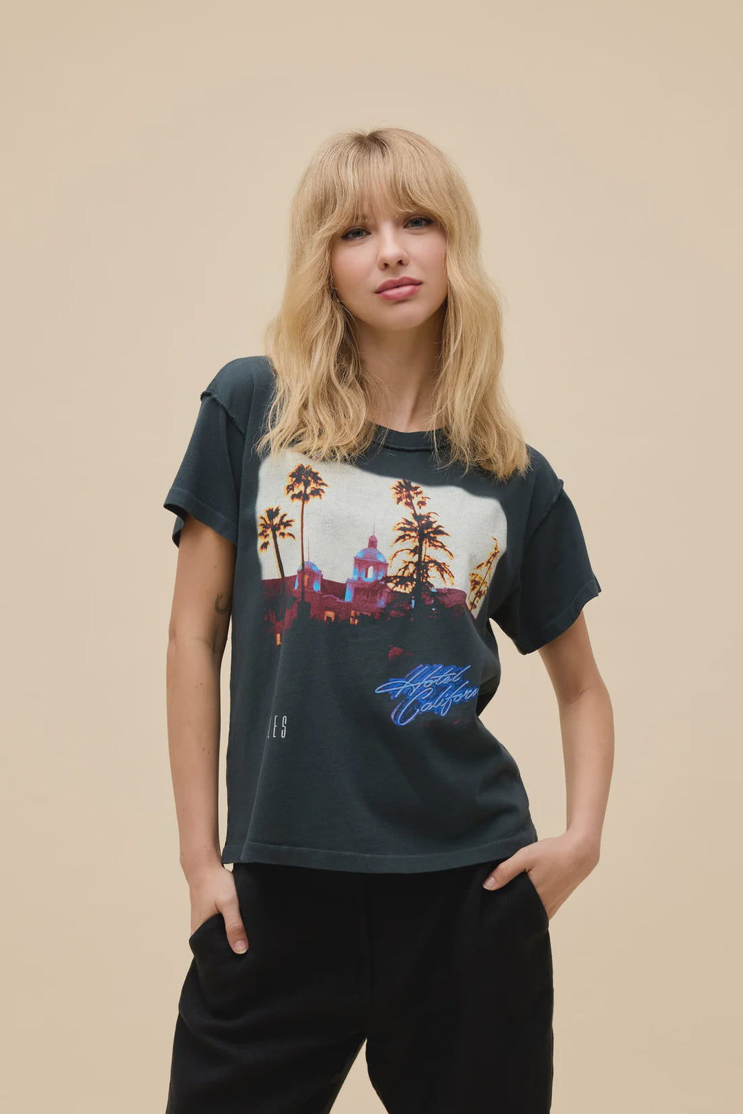 A woman with blonde, wavy hair and bangs wears the Zertz EAGLES HOTEL CALIFORNIA REVERSE GF TEE, which features palm trees and buildings graphics. She poses relaxedly against a beige background with one hand in her pocket.