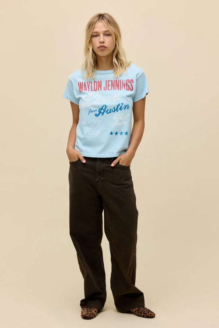 A woman with blonde hair wears a Zertz WAYLON JENNINGS LIVE FROM ATX REVERSE GF TEE in light blue, paired with loose brown pants and leopard-print shoes, standing against a plain beige background.