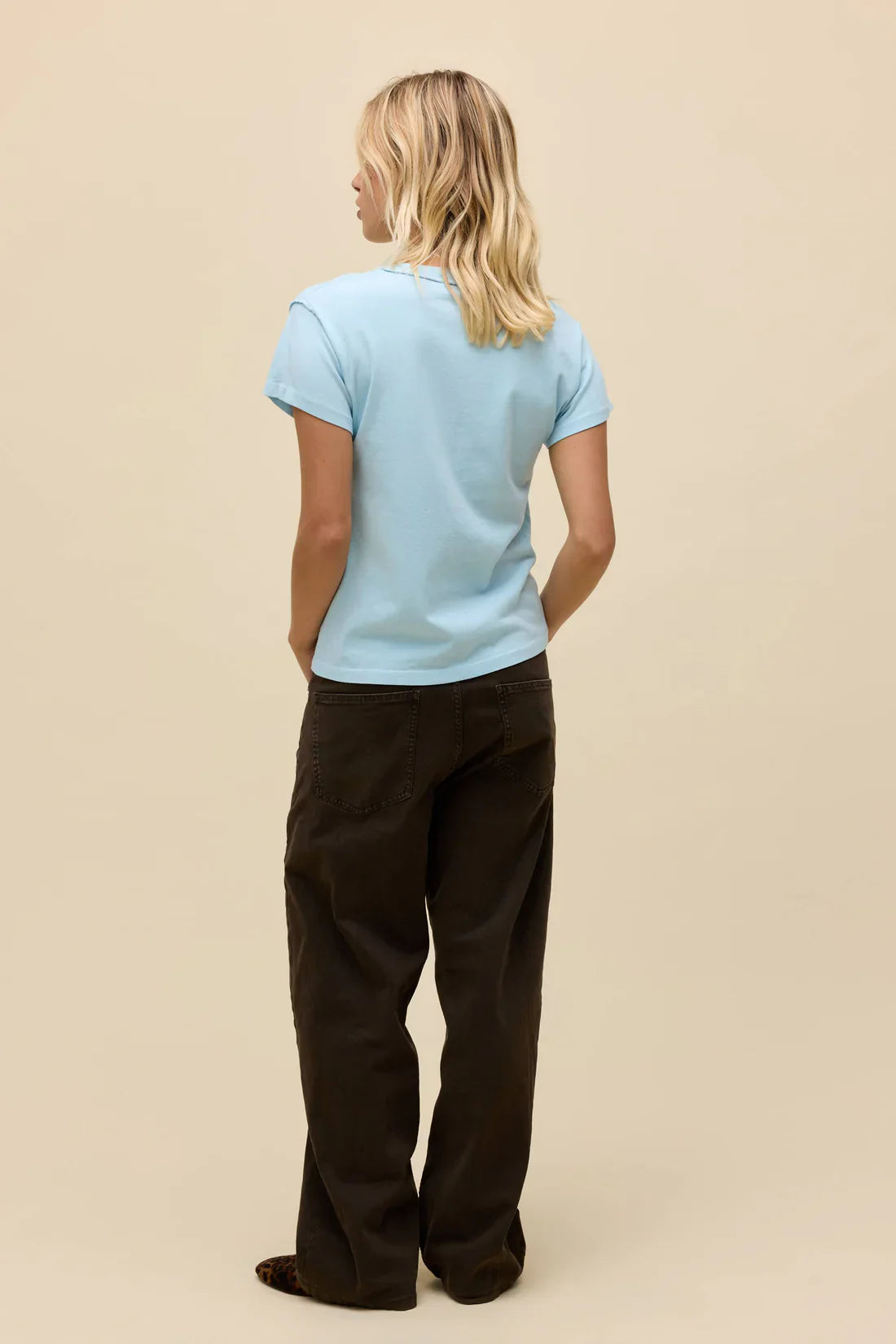 A person with shoulder-length blonde hair stands facing away, wearing the Zertz WAYLON JENNINGS LIVE FROM ATX REVERSE GF TEE in light blue and loose dark brown pants, channeling an Outlaw style against a plain beige background.