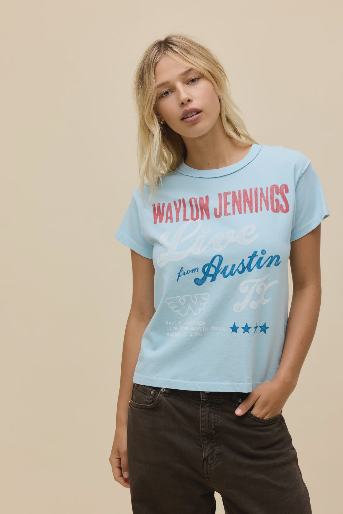 A blonde woman wears the Zertz WAYLON JENNINGS LIVE FROM ATX REVERSE GF TEE, a light blue graphic shirt with the Flying W logo and "Live from Austin, TX," standing against a plain beige background with one hand in her pocket.