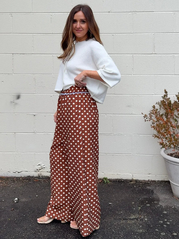 A woman stands before a white brick wall, wearing a white long-sleeve top and Zertz POLKA DOT PANTS—brown, high-waisted wide-leg pants with playful white dots. A potted plant is visible to her right.