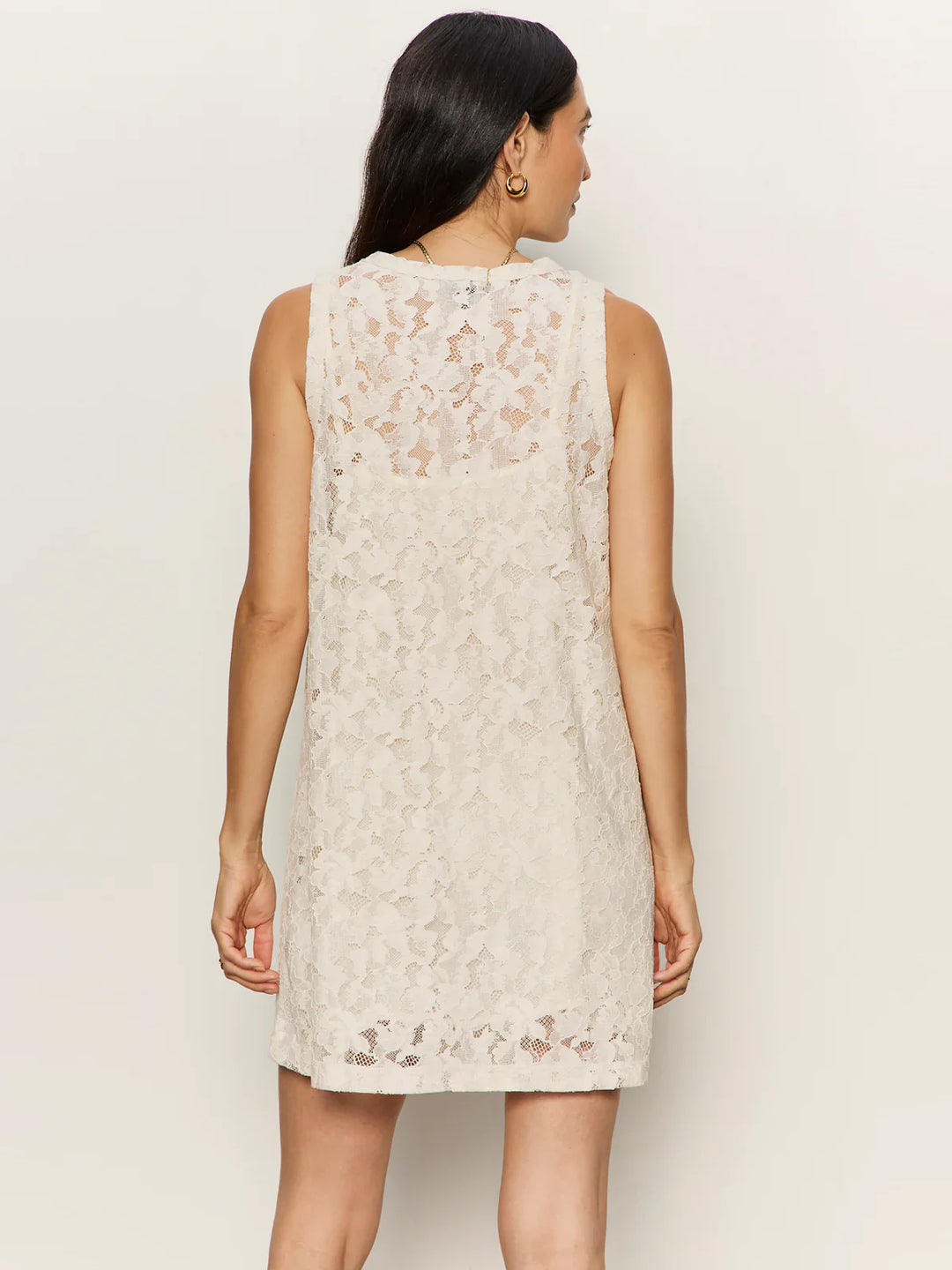 A woman with long dark hair is shown from the back wearing the Zertz SUNDAY BEST LACE TANK DRESS, a cream sleeveless dress with a sheer lace overlay, visible detachable slip, and knee-length hem, set against a plain light background.
