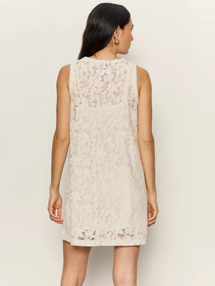 A woman with long dark hair is shown from the back wearing the Zertz SUNDAY BEST LACE TANK DRESS, a cream sleeveless dress with a sheer lace overlay, visible detachable slip, and knee-length hem, set against a plain light background.