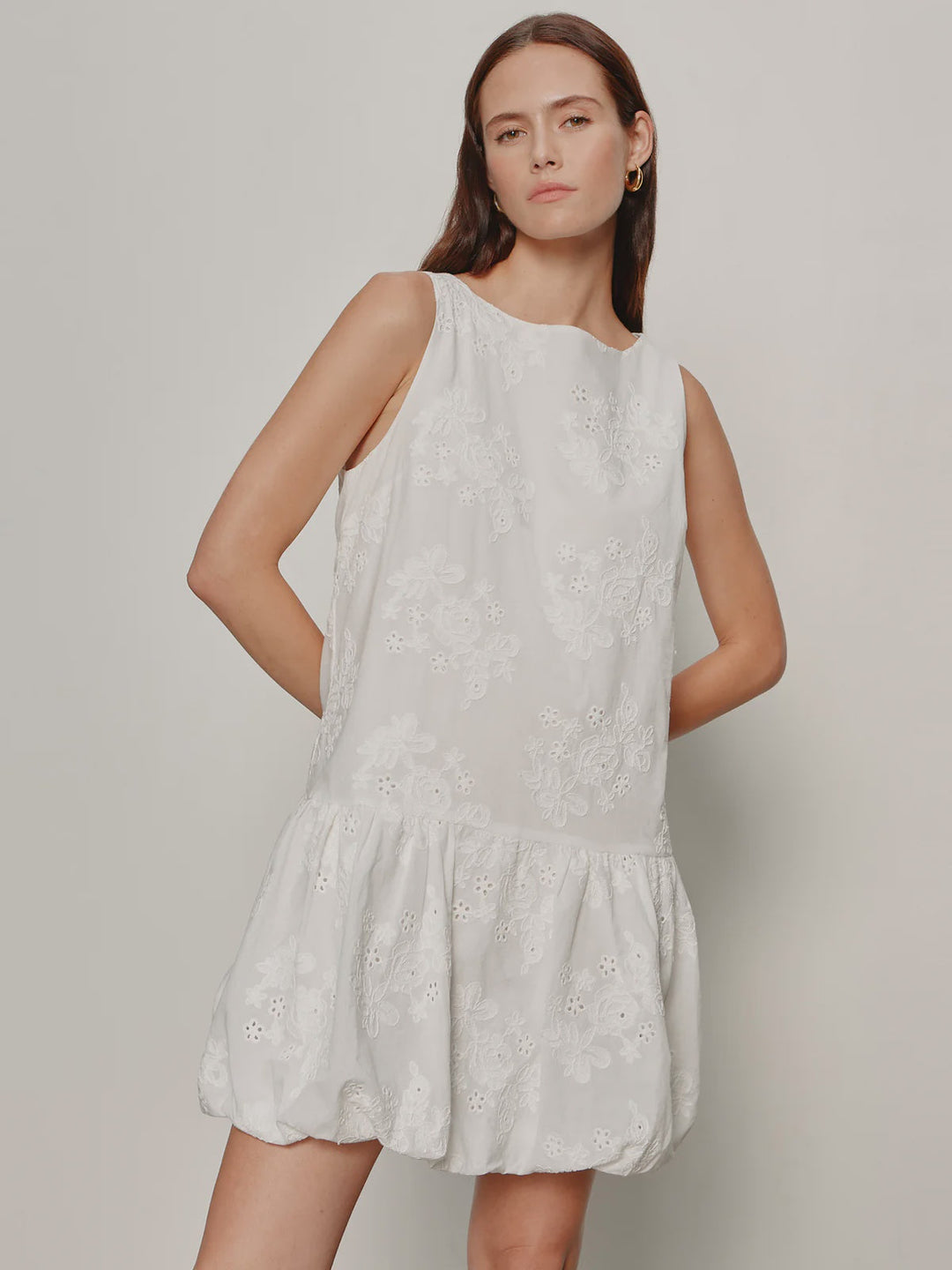 A woman with straight brown hair and a neutral expression stands against a plain background, wearing the Zertz GARDEN EYELET BUBBLE DRESS—a mini dress with floral embroidery, crew neck, relaxed fit, and her arms behind her back.