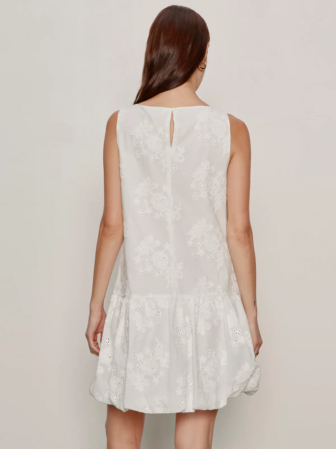 A woman with long brown hair, seen from behind, wears the Zertz GARDEN EYELET BUBBLE DRESS—a relaxed fit, sleeveless white dress with floral embroidery, ruffled hem, crew neck, and a small button closure at the upper back.