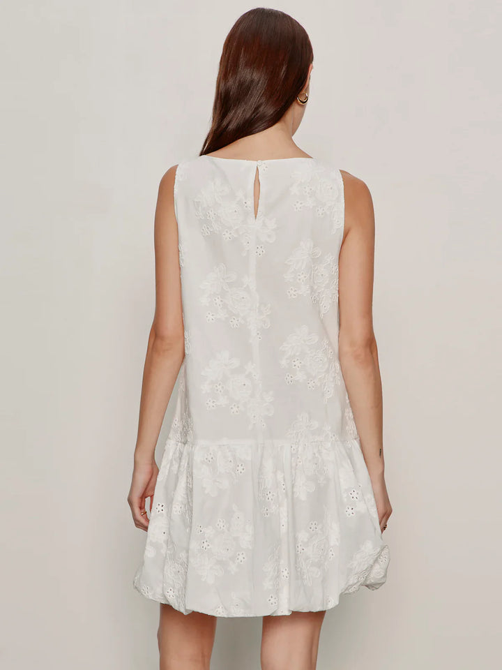 A woman with long brown hair, seen from behind, wears the Zertz GARDEN EYELET BUBBLE DRESS—a relaxed fit, sleeveless white dress with floral embroidery, ruffled hem, crew neck, and a small button closure at the upper back.