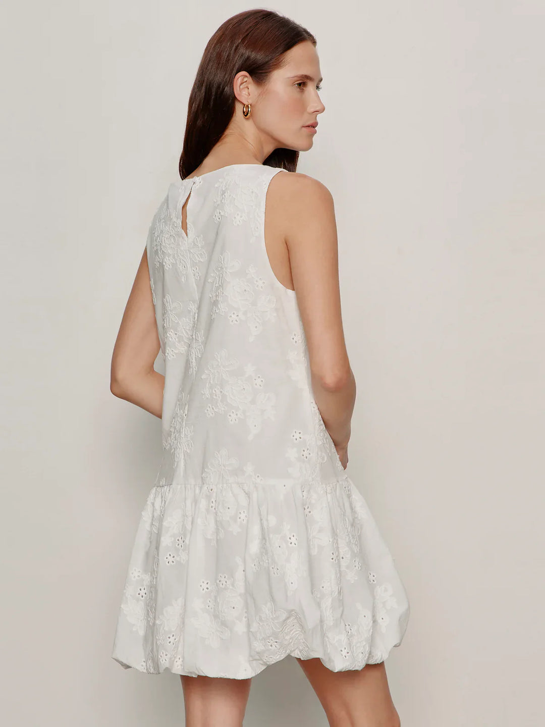A woman with long brown hair models the Zertz GARDEN EYELET BUBBLE DRESS— a relaxed fit, sleeveless white dress with floral embroidery and a ruffled hem—posing with her back to the camera against a light background.