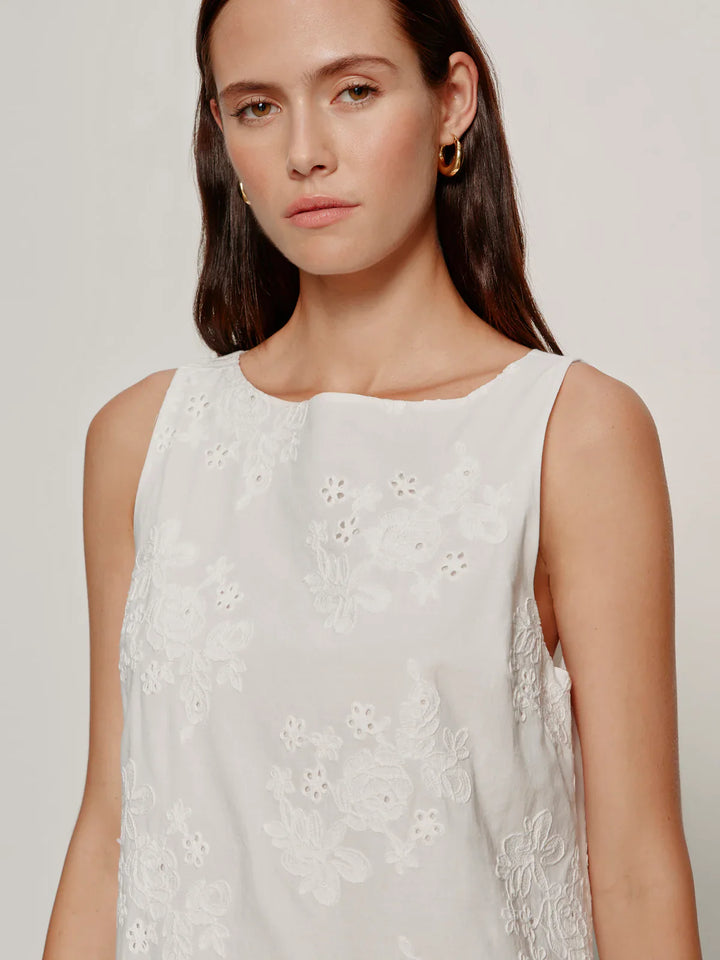 A woman with long brown hair wears the Zertz GARDEN EYELET BUBBLE DRESS, a sleeveless white piece with floral embroidery and a relaxed fit, styled with gold hoop earrings against a plain light background.