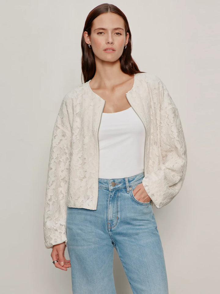 A woman with long brown hair wears the Zertz LEILA LACE JACKET, featuring an all-over lace texture and relaxed fit, over a white tank top and light blue high-waisted jeans, standing against a plain light background.