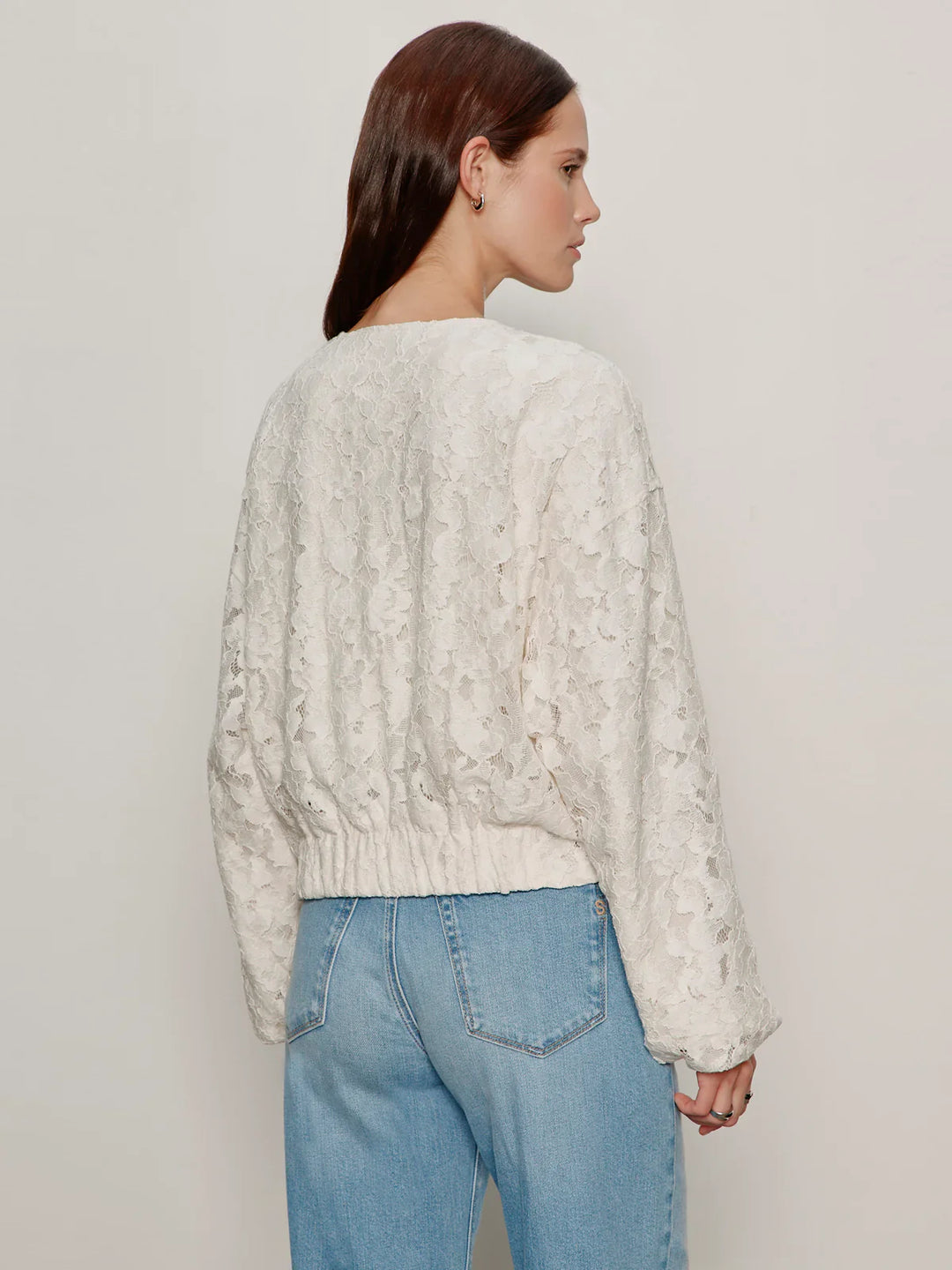 A woman with straight brown hair faces away, wearing the Zertz LEILA LACE JACKET—an all-over lace, long-sleeve off-white top with a relaxed fit—paired with light blue high-waisted jeans against a neutral background.