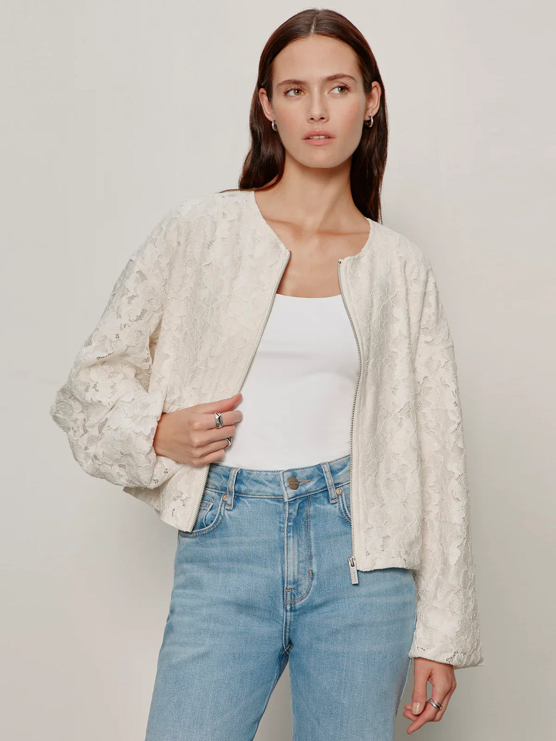 A woman wears the Zertz LEILA LACE JACKET with bubble sleeves over a white tank top and light blue jeans, standing against a plain light background and gazing slightly to the side with a neutral expression.