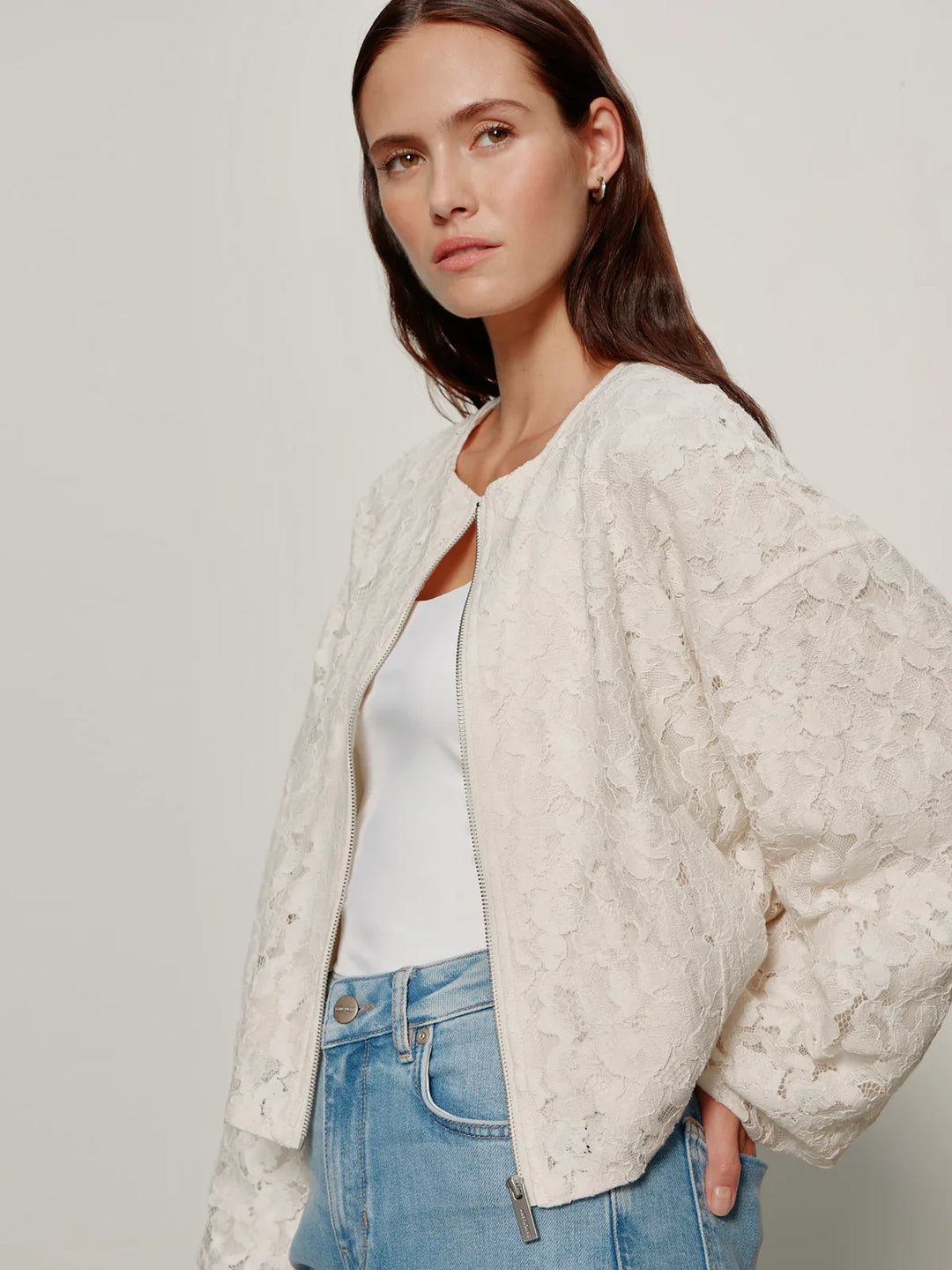 A woman models the Zertz LEILA LACE JACKET over a white top and light blue jeans, standing against a plain light background and looking slightly to the side with a neutral expression.