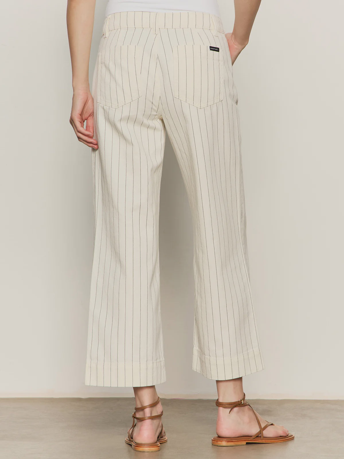 A person is seen from the back wearing Zertz’s THE MARINE PANT in cream pinstripe with wide cropped legs, paired with flat brown sandals, standing on a light-colored floor.