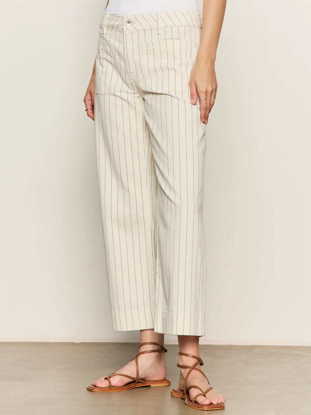 A person wearing Zertz’s THE MARINE PANT in off-white pinstripes, styled with a white top and brown strappy sandals, stands against a plain background.
