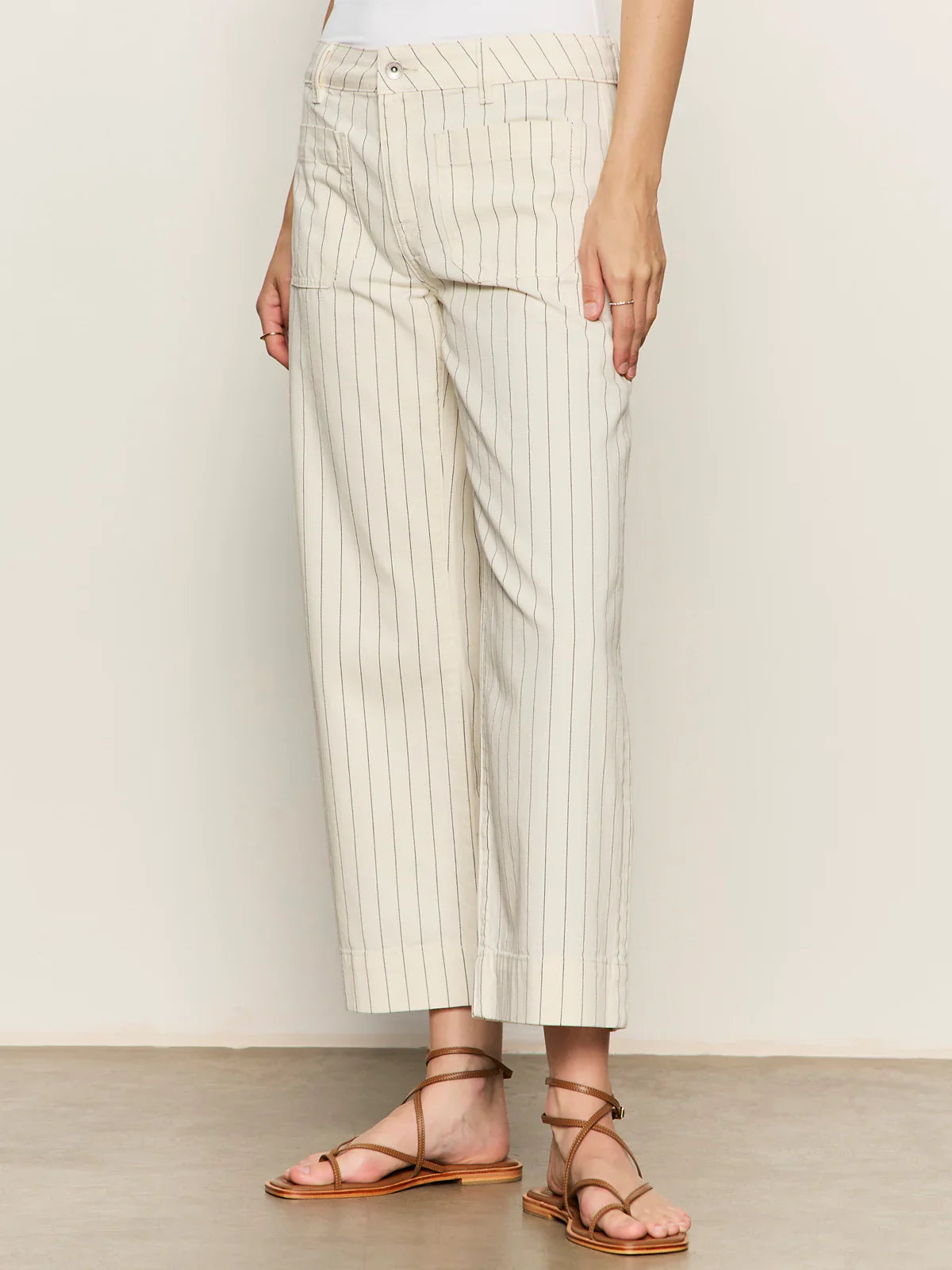 A person wearing Zertz’s THE MARINE PANT in off-white pinstripes, styled with a white top and brown strappy sandals, stands against a plain background.