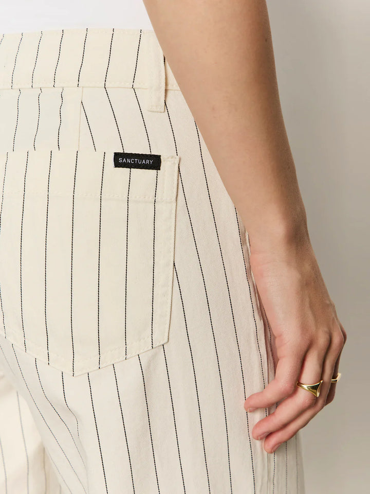 Close-up of a person wearing THE MARINE PANT by Zertz—cream pinstripe cropped wide leg pants with thin black stripes, visible back pocket, and a hand with a gold ring near the hip.