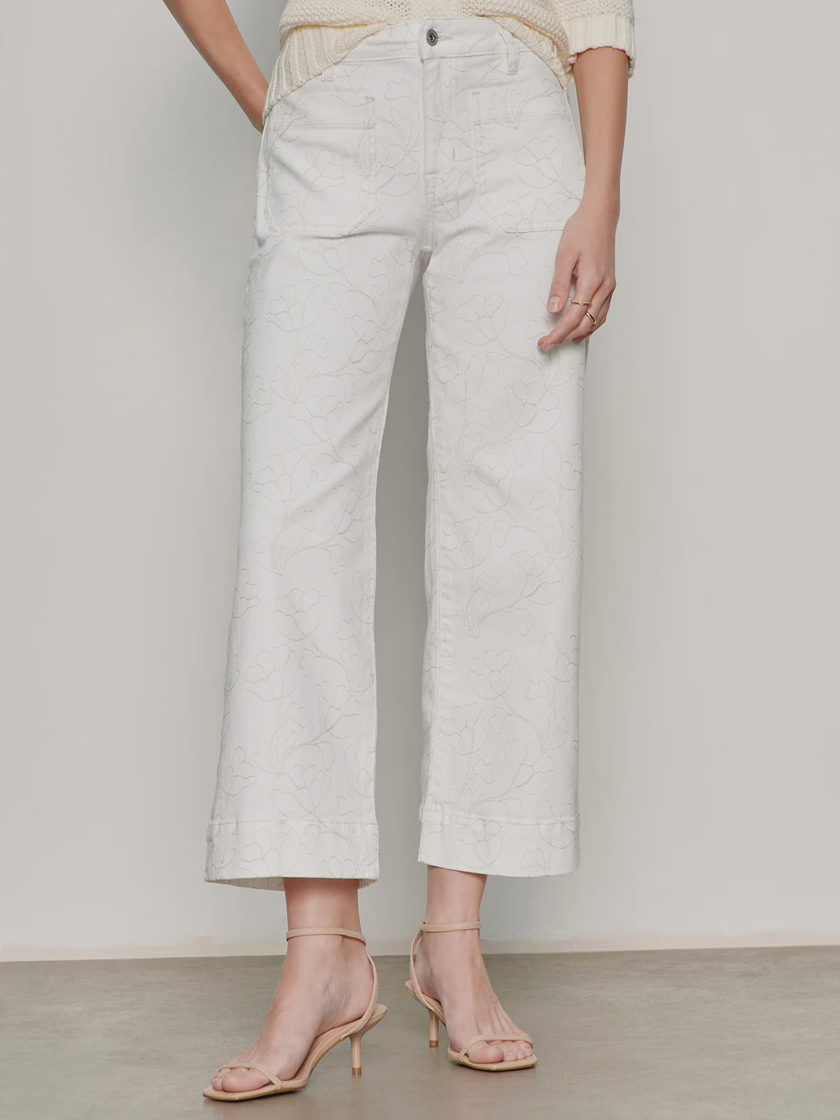 A person models Zertz's THE MARINE CROP: relaxed-fit, white cropped wide-leg pants with a subtle floral print and front patch pockets, paired with light beige high-heeled sandals against a plain background.