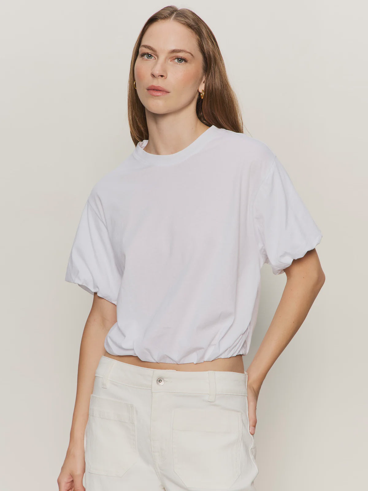 A woman with long brown hair wears the Zertz PLEAT BUBBLE TEE and high-waisted white relaxed-fit pants, standing against a plain, light background with a neutral expression.