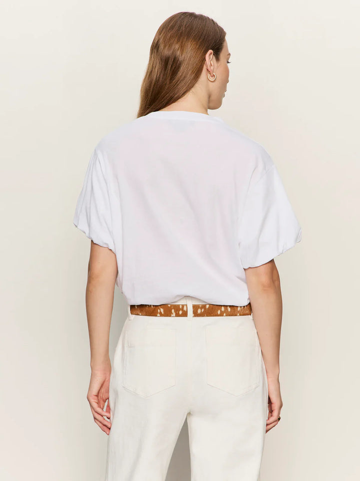 A person with long brown hair wears the Zertz PLEAT BUBBLE TEE, a relaxed fit white pleated short sleeve top tucked into high-waisted off-white pants with a tan patterned belt, standing back to camera against a neutral background.