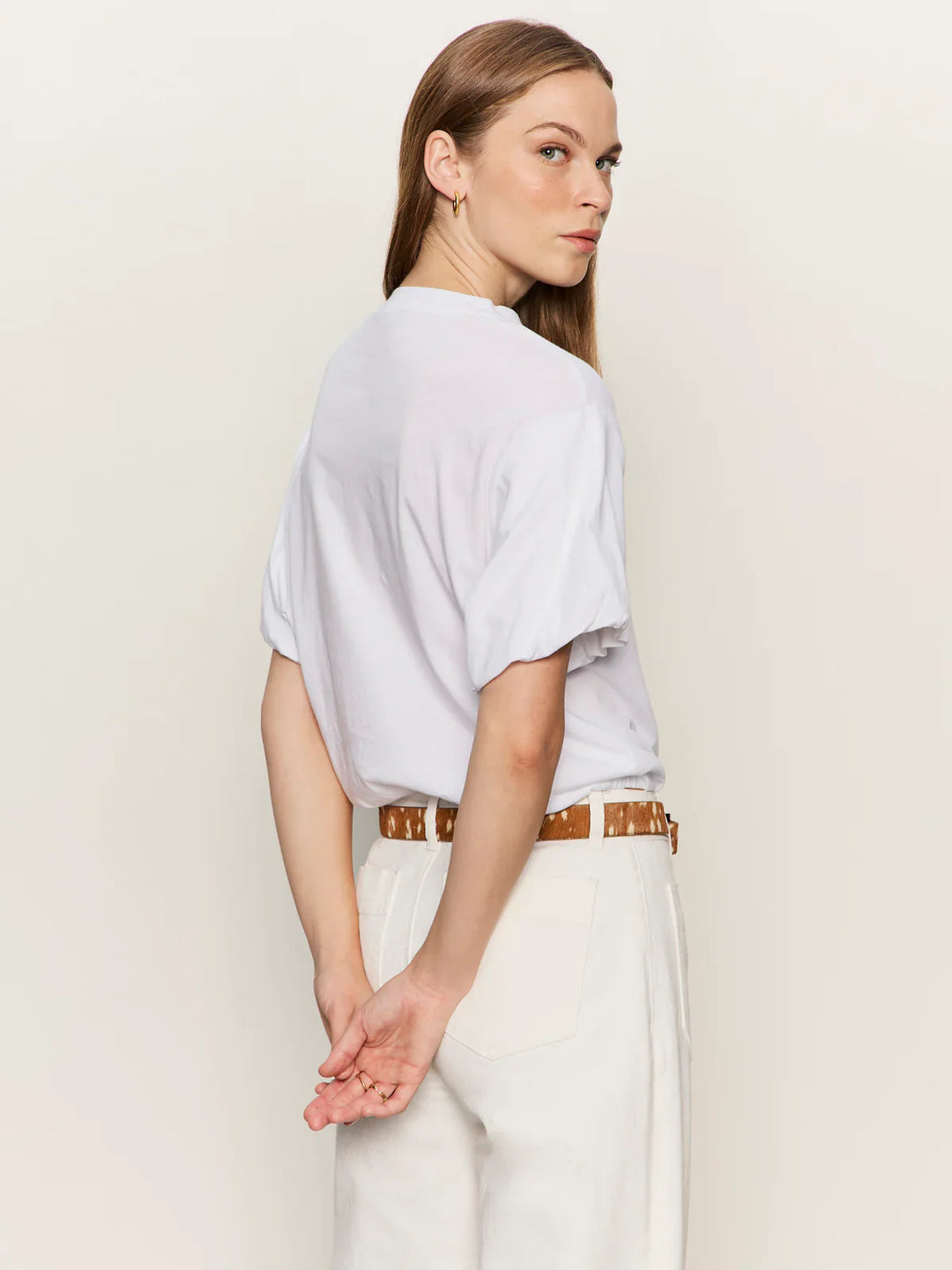 A woman with light brown hair models the Zertz PLEAT BUBBLE TEE in white, paired with white pants and a tan belt. She stands with hands clasped behind her back, looking over her shoulder against a plain light background.