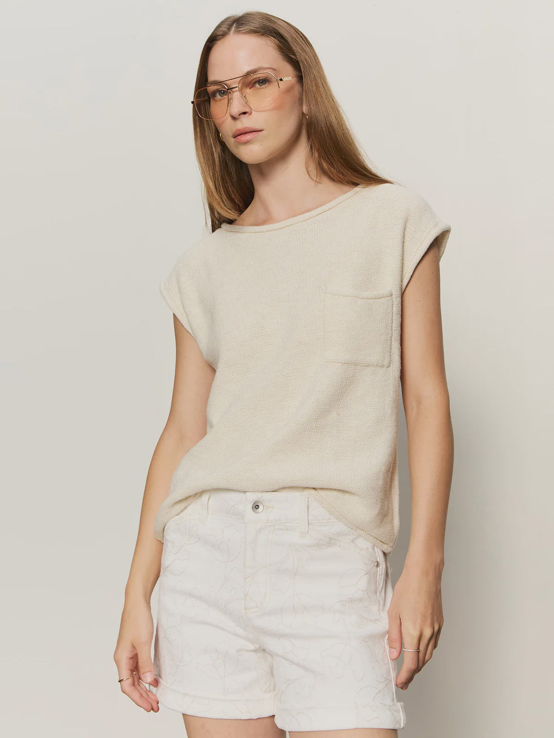 A woman with long brown hair and clear glasses wears the Zertz EASY SWEATER TEE in COLLAGEN, a beige crew neck short sleeve knit top with front pocket and relaxed fit, paired with white patterned shorts, standing against a plain light background.