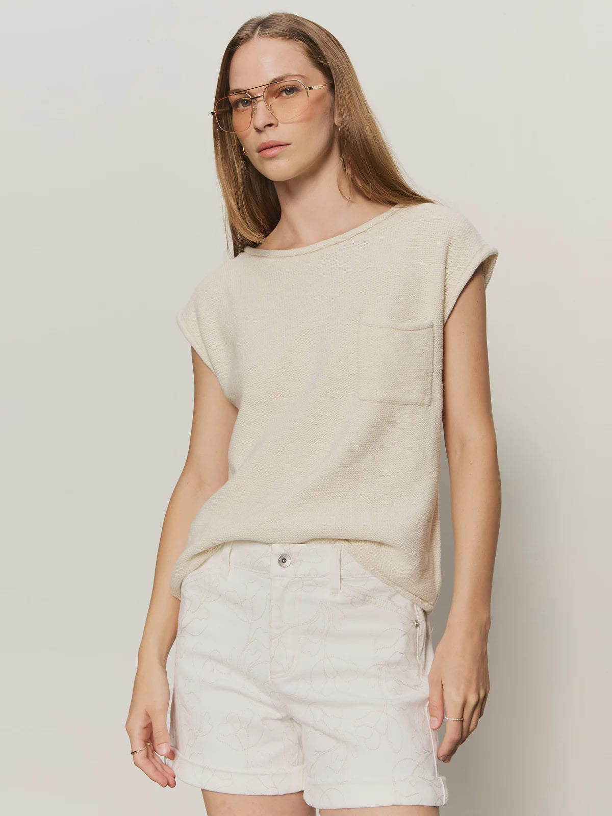 A woman with long brown hair and clear glasses wears the Zertz EASY SWEATER TEE in COLLAGEN, a beige crew neck short sleeve knit top with front pocket and relaxed fit, paired with white patterned shorts, standing against a plain light background.